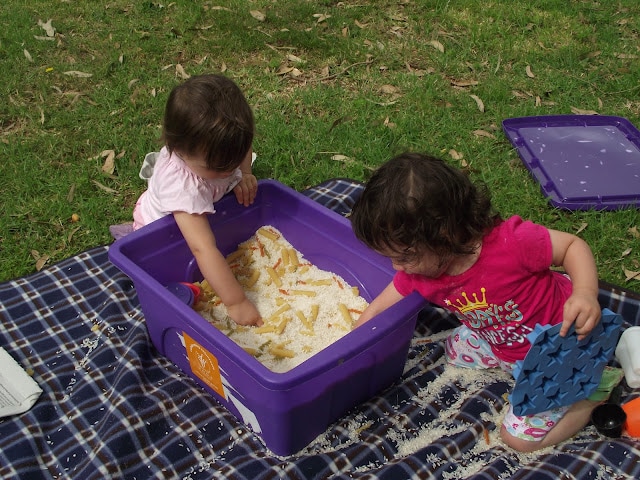 2 toddlers digging into a deep rice and pasta sensory tub, scooping and pouring. 