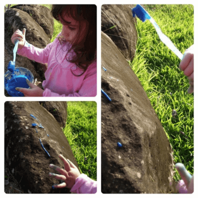 toddler painting outside using a toothbrush on rocks with blue paint in outdoors